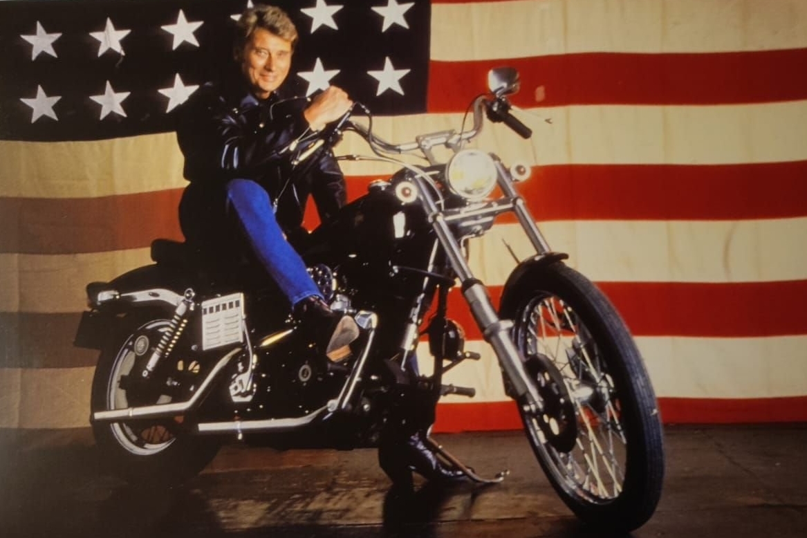 photo de Johnny sur une Harley Davidson lors de sa jeunesse avec un drapeau Américain en fond