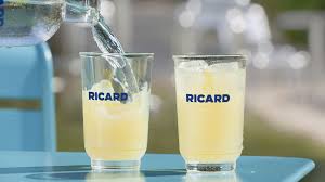 L'image est un photo de deux verre de Ricard, l'un est rempli et l'autre se fait servir. Chaque verre a plusieurs glaçons et il est écrit Ricard dessus. Le fond  est flouté mais on devine que c'est un endroit dans une rue